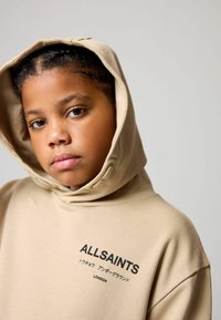 Beżowa bluza z kapturem wykonana z miękkiego materiału. Na przedzie znajduje się czarne logo "ALLSAINTS". Prosty design z luźnym krojem.