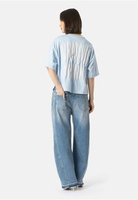 Žena stojící s zády, oblečená v světle modrém oversized tričku s nápisem "CO COKOLIV SI MYSLÍTE" a volných modrých džínách s podpatky.