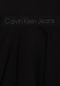 Svart bomullsstoff med en jevn tekstur, som har et horisontalt bånd med "Calvin Klein Jeans" i grå brodering.