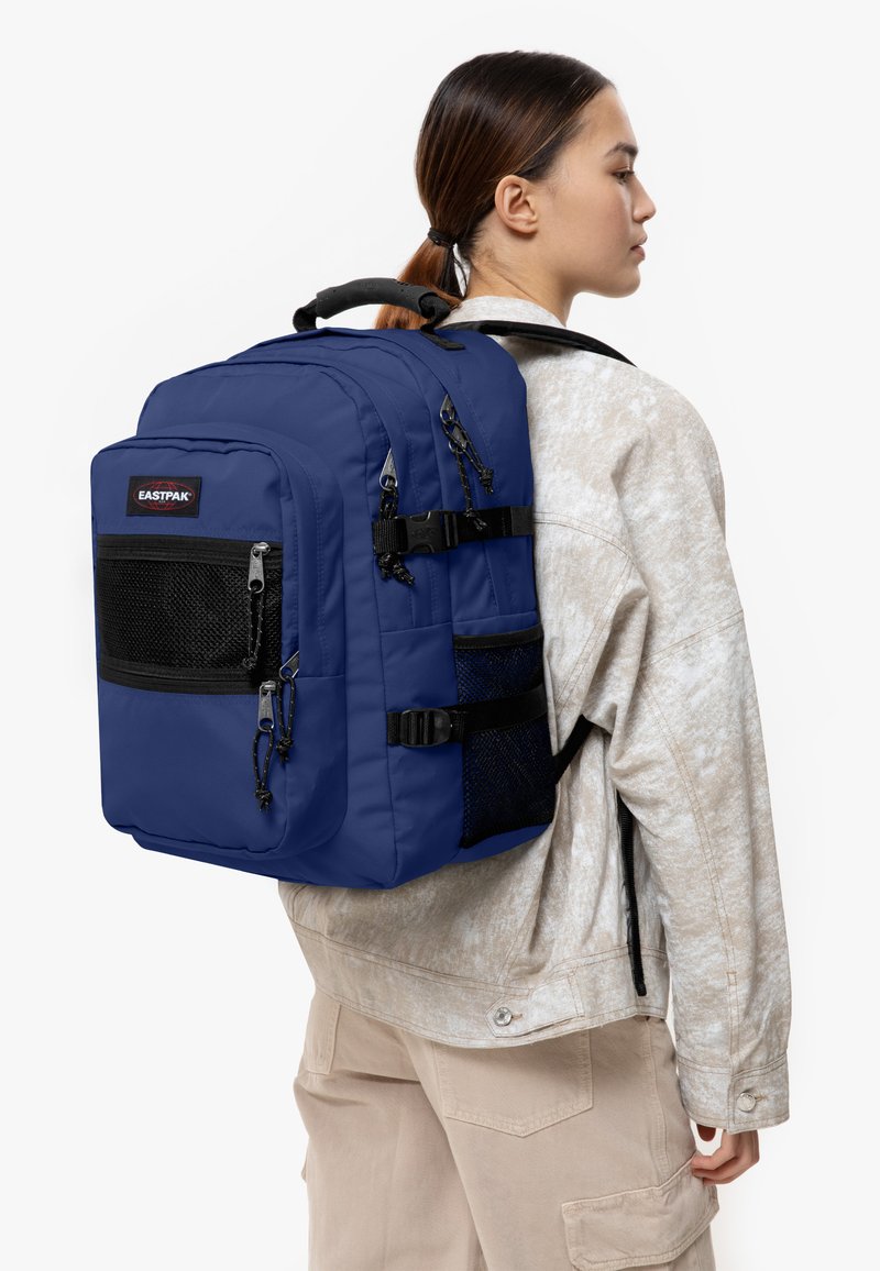Marininblå Eastpak-ryggsäck med flera dragkedjefickor, nät-accenter och justerbara remmar, med ett vadderat handtag på toppen.