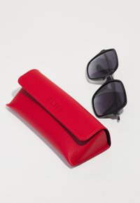 Etui pentru ochelari din piele roșie, cu un finisaj texturat și închidere magnetică, asociat cu ochelari de soare mari, negri, cu lentile închise la culoare.