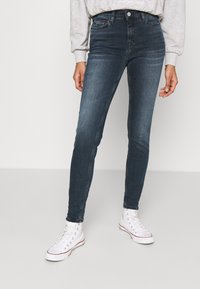 Person som bär mörkblå slitna jeans och vita höga sneakers, står mot en enfärgad vit bakgrund.