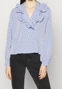 Blusa azzurro chiaro con pois neri, colletto arricciato, chiusura a bottoni e maniche lunghe con polsini elasticizzati. Indossata con pantaloni neri.