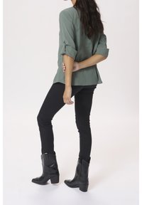 Camicia verde leggera con bottoni e maniche arrotolate; abbinata a jeans neri slim-fit e stivali da cowboy neri con tacco basso.