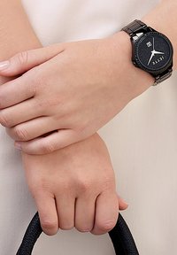 Runder schwarze Uhr mit strukturiertem Ziffernblatt und silbernen Zeigern, an einem Metallarmband. Hand ruht auf einem schwarzen Taschenhenkel, vor einem hellen Hintergrund.