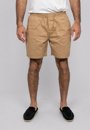 Homme portant un short beige à cordon, un t-shirt blanc, des chaussures noires sans lacets, debout devant un fond gris clair uni.