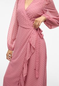 Abito avvolgente rosa con pois testurizzati, dettagli festonati lungo la gonna, maniche a palloncino lunghe e una cintura legata in vita.