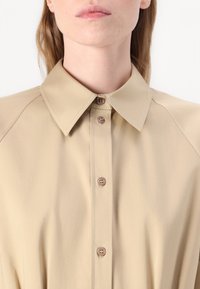 Camicia beige con colletto e chiusura a bottoni frontale, realizzata in un tessuto morbido e con dettagli di cucitura discreti lungo il colletto e le spalle.