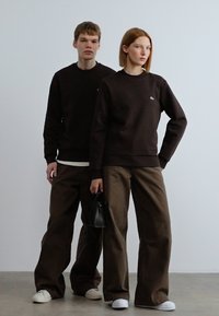 Δύο μοντέλα φορούν σκούρα καφέ φούτερ και καφέ παντελόνια με φαρδύ πόδι. Το γυναικείο μοντέλο κρατά μικρή μαύρη τσάντα χειρός. Και οι δύο φορούν λευκά παπούτσια.