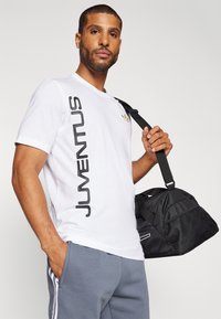 Camiseta blanca con texto negro "JUVENTUS" en el lateral, logo dorado en el pecho, combinada con pantalones grises y una bolsa de deporte negra con detalles en malla.