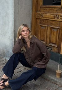 Camicia marrone con bottoni e dettagli in pizzo, abbinata a jeans scuri a gamba larga. Scenografia con un muro testurizzato e una porta di legno; caffè ghiacciato nei dintorni.