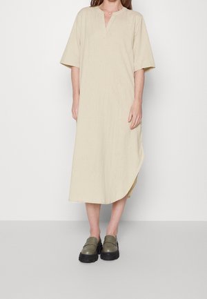 Kobieta ubrana w luźną, beżową sukienkę sięgającą połowy łydki z rękawami do łokci oraz oliwkowozielone masywne mokasyny, stojąca na jednolitym tle.