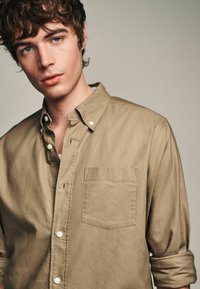 Camisa de botones beige con mangas largas, bolsillo en el pecho y puños doblados. La tela tiene una textura suave con un ligero brillo.
