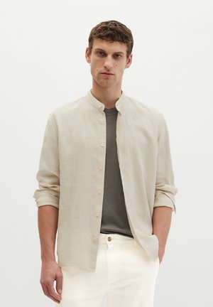 Młody mężczyzna z krótkimi, kręconymi włosami ubrany w beżową koszulę na guziki na szarej koszulce i białe spodnie, stojący na jednolitym tle.