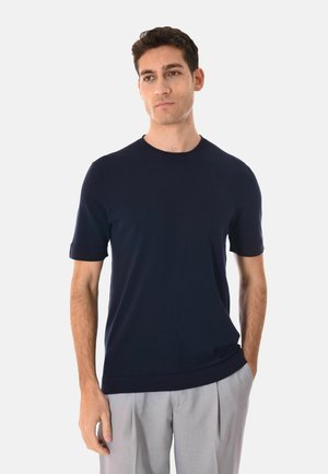 Uomo che indossa una maglia a maniche corte blu navy e pantaloni plissettati grigio chiaro, in piedi con una mano in tasca.