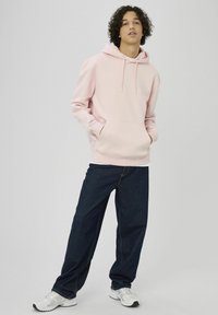 Sweat à capuche rose clair avec une poche avant et des cordons de serrage, associé à un jean en denim bleu foncé. Le mannequin porte des chaussures de sport blanches.