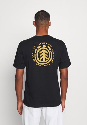 Mann trägt schwarzes T-Shirt mit gelber kreisförmiger Grafik und den Worten "Earth," "Fire," "Wind," "Water," und "Juno", von hinten gesehen.
