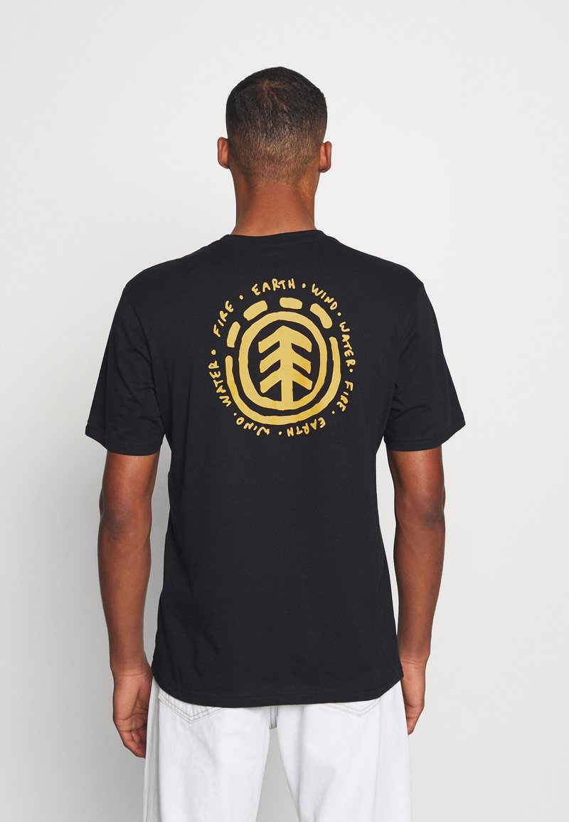 Homme portant un T-shirt noir avec un motif circulaire jaune et les mots "Earth," "Fire," "Wind," "Water," et "Juno" vu de dos.