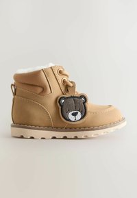 Beige Kinderwinterstiefel mit weißem Fleece-Futter und einem bärenförmigen Schlüsselanhänger, der an den Schnürsenkeln hängt, vor einem neutralen Hintergrund.