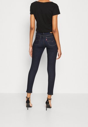 Mujer con vaqueros pitillo azul oscuro y camiseta negra de manga corta, de pie y de espaldas, con sandalias negras de tacón sobre fondo blanco.