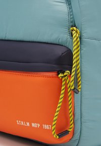 Primo piano di uno zaino multicolore con una tasca arancione, tiranti dello zip in corda gialla e nera e testo stampato sulla tasca.