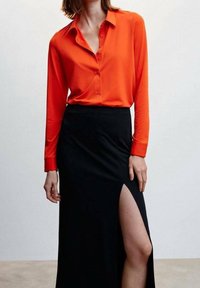 Blouse rouge à manches longues avec fermeture à boutons, associée à une jupe noire avec une fente haute sur la cuisse. Tissu lisse avec un design uni.