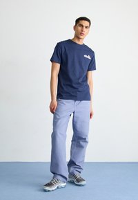 T-shirt en coton marine avec un petit logo, associé à un pantalon droit bleu clair en tissu doux. Les chaussures présentent un design gris avec des accents transparents.