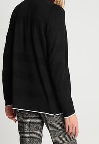 Czarny dziergany kardigan z poziomymi paskami, charakteryzujący się luźnym krojem, długimi rękawami i białym wykończeniem u dołu. Widoczne spodnie w kratę.