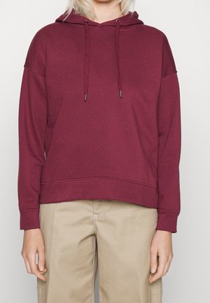 Personne portant un sweat à capuche uni marron foncé avec des cordons et un pantalon beige, debout sur un fond blanc uni.