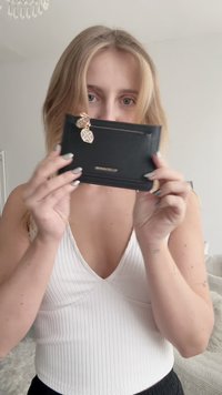 Portafoglio in pelle nera con superficie lavorata, caratterizzato da una zip dorata e un ciondolo decorativo dorato. Forma rettangolare compatta.