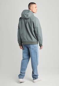 Light gray hooded jacket with a smooth texture, elastic cuffs, and a cropped fit, paired with loose blue jeans and white sneakers.