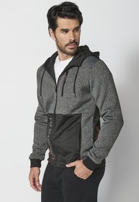 Sudadera con cremallera en gris y negro con un cuerpo texturizado, panel liso negro, capucha ajustable y acentos naranjas en los laterales.