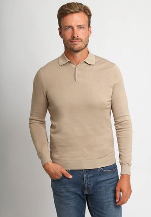 Man met kort blond haar die een beige lange mouwen polo en blauwe spijkerbroek draagt, staand met één hand in de zak tegen een witte achtergrond.