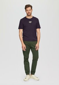 Dunkelviolettes T-Shirt mit Rundhalsausschnitt und kleinem Etikett auf der Brust, kombiniert mit grünen schmalen Hosen und cremigen Sneakers, vor einem hellen Hintergrund stehend.