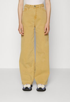 Osoba nosząca spodnie w kolorze musztardowym, z wysokim stanem i szerokimi nogawkami, z przednimi kieszeniami oraz białe trampki z fioletowymi i czarnymi akcentami.