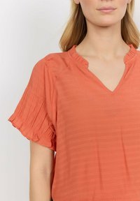 Short-sleeve, V-neck blouse in coral, made of textured fabric with a subtle stripe pattern. Features ruffled edges on sleeves.