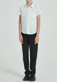 Chemise blanche à manches courtes avec une poche poitrine, associée à un pantalon noir et des chaussures noires, portée par une personne debout.