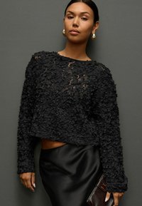 Zwarte cropped trui met gestructureerde bloemenapplicaties, lange mouwen, ronde halslijn, gecombineerd met een strakke zwarte satijnen rok en een metallic clutch.