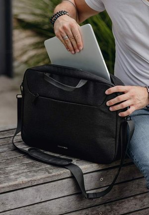 Černá látková taška na notebook s horním držadlem a nastavitelným ramenním popruhem, která má přední kapsu a elegantní design, umístěná na dřevěném povrchu.