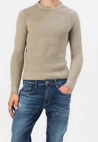 Beżowy sweter z dzianiny z prążkowanym okrągłym dekoltem i długimi rękawami, zestawiony z niebieskimi dżinsami o klasycznym, pięciokieszeniowym kroju.