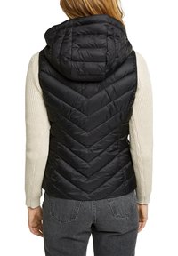 Personne portant une veste matelassée noire avec capuche sur un pull en tricot crème et un jean gris foncé, vue de dos.