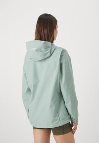 Lys grønn vanntett jakke med hette, med elastiske mansjetter og en subtil logo på baksiden. Glatt tekstur med minimalistisk design.