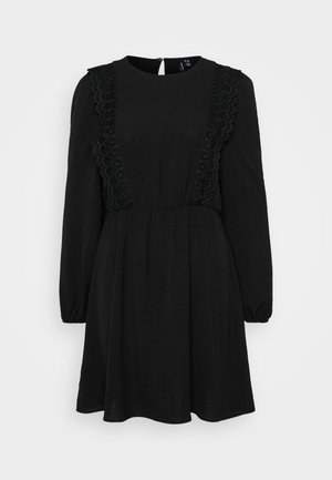 Robe noire à manches longues et col rond. Présente des détails en dentelle texturée sur les côtés et une taille cintrée. Fermeture au dos.