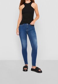 Top negro de canalé sin mangas con cuello alto, combinado con jeans ajustados azules y sandalias de slide negras de trenzado con suela gruesa.