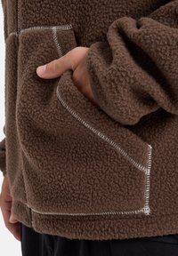 Chaqueta de forro polar marrón con un acabado texturizado, costuras blancas contrastantes y bolsillos frontales a cada lado.