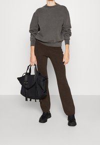Sweatshirt oversized gris charbon, pantalons ajustés marron et sac fourre-tout noir avec des sangles. Porté avec des baskets noires en tissu texturé.