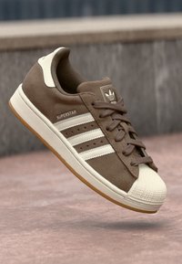 Sneaker in suede marrone con accenti crema, caratterizzato da tre strisce bianche, punta in gomma e suola in gomma marrone. Modello: Superstar.