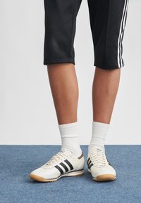 Witte sportschoenen met zwarte strepen, een bruine zool en gestructureerd leer. Draagt witte sokken en zwarte korte broek met witte strepen.