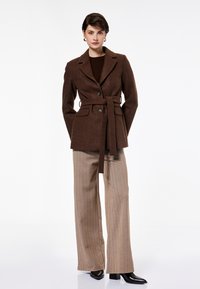 Blazer de mezcla de lana marrón con cintura ceñida con cinturón, dos botones y solapas en los bolsillos. Combinado con pantalones anchos de rayas finas beige y botas negras de tacón.