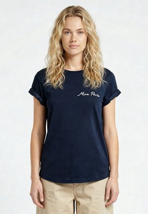Jonge vrouw met lang golvend blond haar die een marineblauw T-shirt draagt met de tekst "Mira Paris" en beige broek, staand tegen een lichte achtergrond.